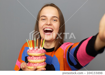 Indoor shot of smiling attractive young adult woman with wearing warm sweater holding donut with candles isolated over gray background, making selfie, being excited. 104133898