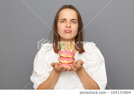 Indoor shot of sad upset woman with brown hair wearing white blouse, looking at camera with pout lips, has no gusts at her birthday party, holding donut with candles isolated over gray background. 104133904