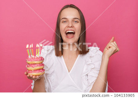 Portrait of overjoyed good looking pretty woman with brown hair wearing white blouse, clenched fist, celebrating her birthday holding donut with candles isolated over pink background. 104133905