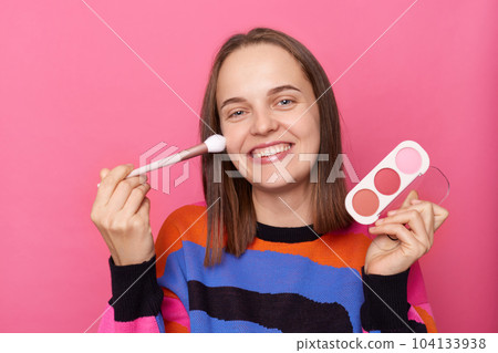 Portrait of smiling delighted woman with dark straight hair wearing jumper standing isolated over pink background, holding palette of eyeshadow in hands, applying powder with brush on her cheek. 104133938
