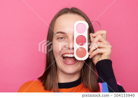 Portrait of young Caucasian woman covering her eye with colorful eyeshadow palette, wearing sweater, doing makeup for dating, posing isolated over pink background. Portrait of young Caucasian woman covering her eye with colorful eyeshadow palette, wearing sweater, doing makeup for dating, posing isolated over pink background. 104133940