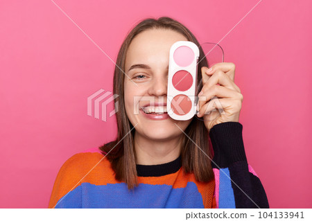 Image of joyful cheerful woman with brown hair wearing sweater, covering her eye with eyeshadow palette, looking at camera with toothy smile, posing isolated over pink background. Image of joyful cheerful woman with brown hair wearing sweater, covering her eye with eyeshadow palette, looking at camera with toothy smile, posing isolated over pink background. 104133941