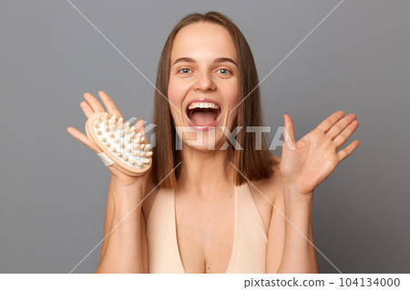 Indoor shot of excited joyful woman uses dry body brush wants to have smooth clean skin, yelling with excitement, poses against gray background, posing with open mouth. Indoor shot of excited joyful woman uses dry body brush wants to have smooth clean skin, yelling with excitement, poses against gray background, posing with open mouth. 104134000