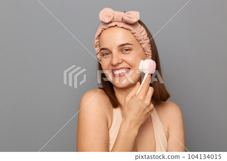 Portrait of emotional woman with hair band with perfectly groomed skin, posing isolated over gray background, doing facial procedures with new brush for washing. 104134015