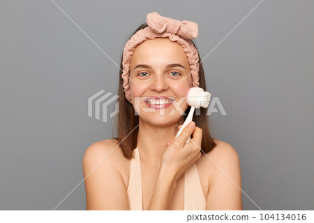 Indoor shot of smiling happy woman standing isolated over gray background, using brush for washing, doing morning routine procedures, looking at camera with happy expression, enjoying skin care. 104134016