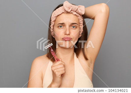 Indoor shot of sad upset woman shaving armpit with pink shaver isolated on gray background, choosing bad variant of epilation, looking at camera with pout lips, being disappointed. 104134021