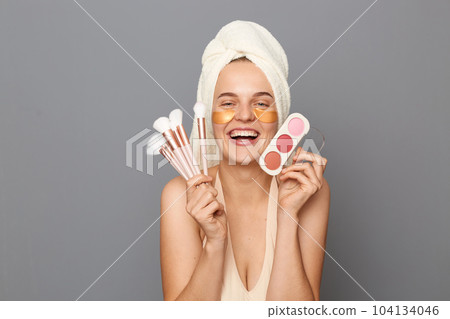 Indoor shot of young adult attractive woman holding shadows for eyelids, posing isolated over gray background, charming girl makes morning beauty treatments and day make-up. 104134046