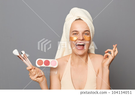 Indoor shot of extremely happy laughing woman in towel on her head, holding eye shadows and brushes in hands, crossing fingers for go luck for good makeup. 104134051