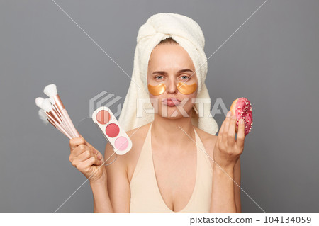Sad upset woman wrapped in towel standing cosmetics brushes and palette of eye shadows and tasty donut, expressing sorrow, can't eat, being on diet, posing isolated over gray background. Sad upset woman wrapped in towel standing cosmetics brushes and palette of eye shadows and tasty donut, expressing sorrow, can't eat, being on diet, posing isolated over gray background. 104134059