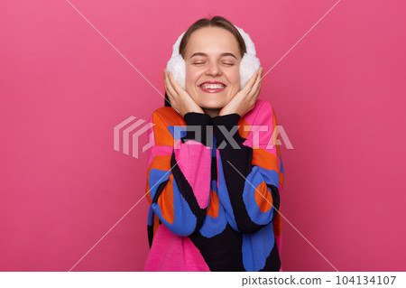 Image of smiling satisfied delighted Caucasian woman wearing warm sweater and fur earmuffs standing isolated over pink background, smiling happily, closed her eyes, enjoying moment. 104134107