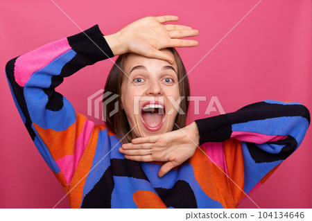 Indoor shot of happy satisfied woman wearing colorful jumper posing isolated over pink background, rejoicing her success, celebrating, screaming with happiness, showing her palms. Indoor shot of happy satisfied woman wearing colorful jumper posing isolated over pink background, rejoicing her success, celebrating, screaming with happiness, showing her palms. 104134646