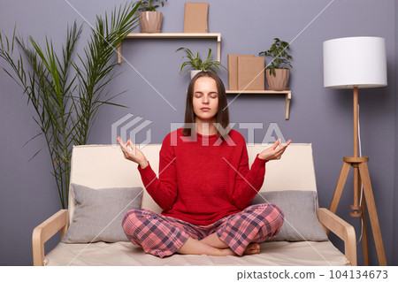 Portrait of calm young adult brown-haired woman wearing red sweater and checkered pants sitting on cough in lotus pose in living room at home, practicing yoga, relaxing. 104134673