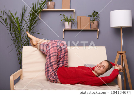 Portrait of happy delighted satisfied woman wearing red sweater and checkered pants lying on sofa in home interior and having rest after hard working day, enjoying weekend. 104134674