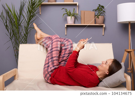 Indoor shot of smiling positive optimistic woman wearing red sweater and checkered pants lying on sofa in home interior and holding mobile phone, chatting with her friends in social networks. 104134675