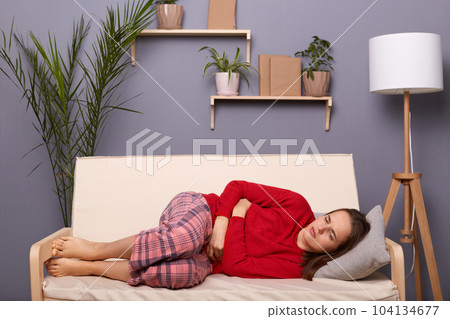 Horizontal shot of sad unhealthy sick woman wearing red sweater and checkered pants lying on sofa in home interior, suffering stomachache, holding her belly, feels period pain. 104134677