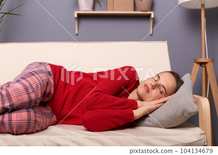 Image of calm tired Caucasian woman wearing red sweater and checkered pants lying on sofa in home interior, keeps closed eyes and sleeping, having nap. Image of calm tired Caucasian woman wearing red sweater and checkered pants lying on sofa in home interior, keeps closed eyes and sleeping, having nap. 104134679