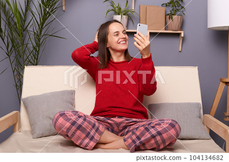 Portrait of smiling positive Caucasian woman wearing red sweater and checkered pants sitting on sofa in home interior, holding smart phone in hands, making selfie or having video call. 104134682