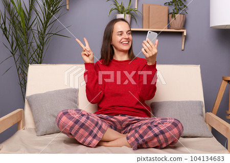 Indoor shot of satisfied young adult woman wearing red sweater and checkered pants sitting on sofa in home interior, holding smart phone, having video call or making selfie, smiling and showing v sign 104134683