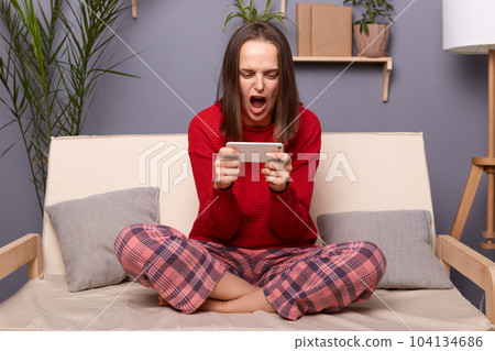 Portrait of sad excited brown-haired woman gamer wearing red sweater and checkered pants sitting on cough in living room at home, playing video game on cell phone, loosing the level. 104134686