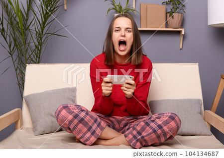 Photo of addicted woman gamer wearing red sweater and checkered pants sitting on sofa in home interior, holding cell phone, lose the level of favorite game, screaming with despair. 104134687