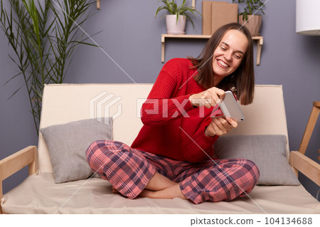 Portrait of excited, extremely woman with dark hair wearing home clothing sitting on sofa in living room, holding mobile phone in hands, playing interesting video game online. Portrait of excited, extremely woman with dark hair wearing home clothing sitting on sofa in living room, holding mobile phone in hands, playing interesting video game online. 104134688