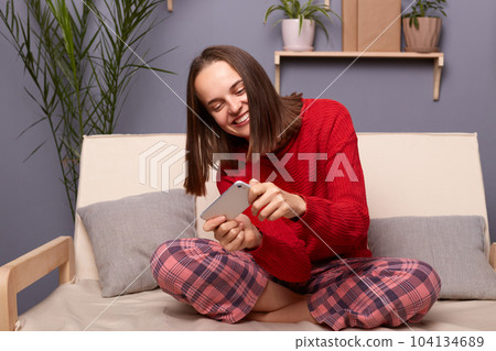 Indoor shot of happy satisfied young adult brown-haired woman blogger wearing red sweater and checkered pants sitting on cough in living room at home, playing video game, rejoices to win the level. 104134689