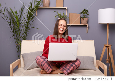 Portrait of positive attractive woman with dark hair wearing home clothing working online on laptop or having video call, sincerely, looking on screen, sitting on sofa in living room. 104134701