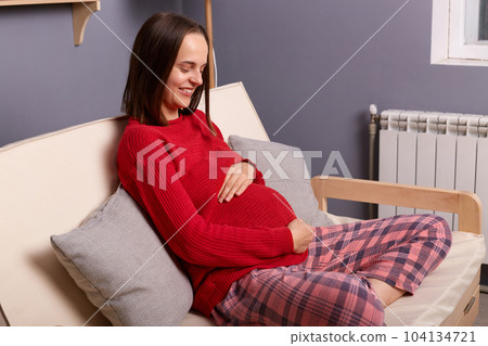 Image of young adult pregnant smiling woman wearing red jumper and pajama pants sitting on sofa in home interior, holding hands on her belly, waiting her baby. Image of young adult pregnant smiling woman wearing red jumper and pajama pants sitting on sofa in home interior, holding hands on her belly, waiting her baby. 104134721