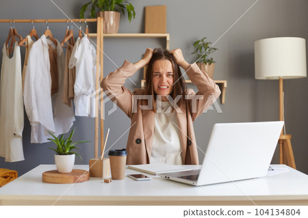 Image of aggressive angry Caucasian brown-haired woman stylist wearing beige jacket sitting at her workplace in front of notebook, screaming, keeping hands on head. 104134804
