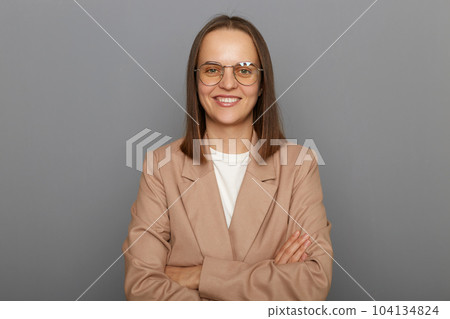 Indoor shot of smiling positive happy woman wearing beige jacket posing isolated over gray background, standing with crossed arms, looking at camera with toothy smile. Indoor shot of smiling positive happy woman wearing beige jacket posing isolated over gray background, standing with crossed arms, looking at camera with toothy smile. 104134824