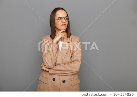 Indoor shot of pensive thoughtful young adult female wearing beige jacket standing isolated over gray background, holding her chin, thinking, looking away. Indoor shot of pensive thoughtful young adult female wearing beige jacket standing isolated over gray background, holding her chin, thinking, looking away. 104134826