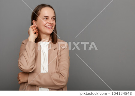 Image of attractive delighted woman with brown hair wearing beige jacket, smiling while looking aside at mockup for advertisement, posing isolated over gray background. 104134848