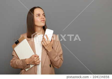 Image of pensive dreaming brown-haired woman wearing jacket holding clipboard with documents and smart phone in hands, standing isolated over gray background. 104134856