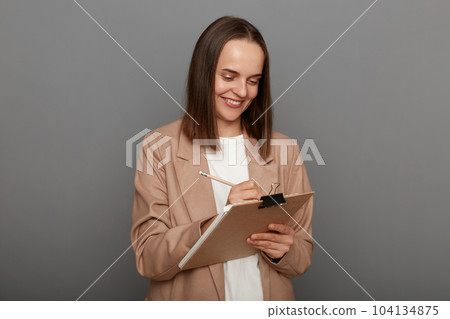 Photo of satisfied delighted woman with brown hair wearing beige jacket posing isolated over gray background, businesswoman writing to do list for working day. 104134875