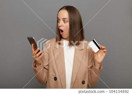 Indoor shot of amazed astonished woman with brown hair wearing beige jacket posing isolated over gray background, holding credit card in hand and using cell phone, being very surprised. Indoor shot of amazed astonished woman with brown hair wearing beige jacket posing isolated over gray background, holding credit card in hand and using cell phone, being very surprised. 104134892