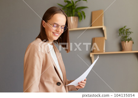 Closeup portrait of smiling woman in glasses with brown hair wearing beige jacket working in office, holding documents in hands, expressing positive emotions, enjoying her work. Closeup portrait of smiling woman in glasses with brown hair wearing beige jacket working in office, holding documents in hands, expressing positive emotions, enjoying her work. 104135054