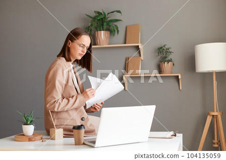 Portrait of young adult attractive businesswoman wearing official style suit holding papers documents and working on laptop, sitting on table in office. Portrait of young adult attractive businesswoman wearing official style suit holding papers documents and working on laptop, sitting on table in office. 104135059