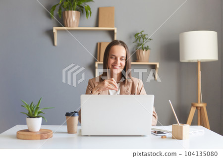 Image of joyful young businesswoman in eyeglasses wearing stylish jacket working online on notebook in home office, looking at computer screen, having video call. 104135080