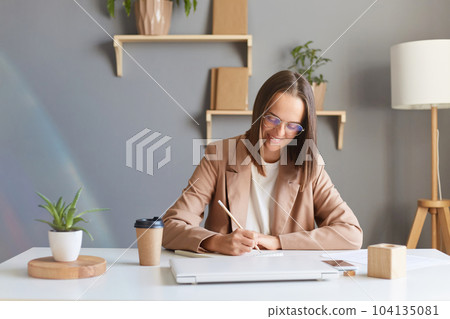 Indoor shot of smiling joyful woman freelancer or designer with brown hair wearing beige jacket working in office, drawing sketches or checking paper documents. 104135081
