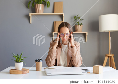Image of tired overworked sick woman with brown hair wearing beige jacket working in office, working hard, feeling fatigue and terrible headache, frowning face, massaging temples. 104135089