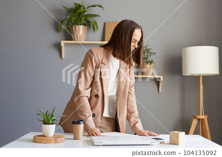 Portrait of brown haired young adult woman with brown hair wearing beige jacket working in office, standing near table, posing at her workplace, looking away. Portrait of brown haired young adult woman with brown hair wearing beige jacket working in office, standing near table, posing at her workplace, looking away. 104135092