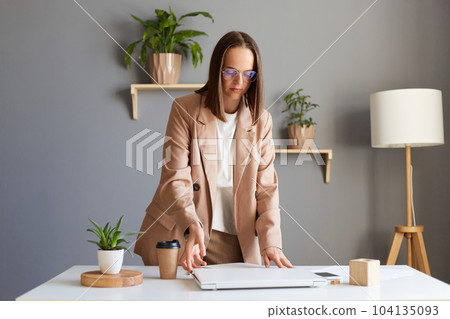 Image of calm serious concentrated attractive young adult Caucasian woman wearing beige jacket posing in office, being ready to start work, open her laptop. Image of calm serious concentrated attractive young adult Caucasian woman wearing beige jacket posing in office, being ready to start work, open her laptop. 104135093