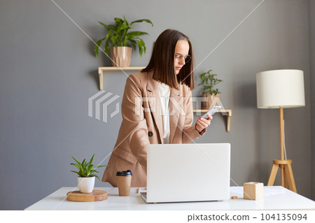 Portrait of dark haired woman freelancer standing near her workplace with notebook and holding smart phone in hands, calling to her partner, dialing number. 104135094