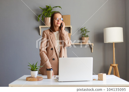 Indoor shot of pretty friendly woman with brown hair wearing beige jacket working online on laptop and talking phone with her partner, looking at camera with calm charming smile. 104135095