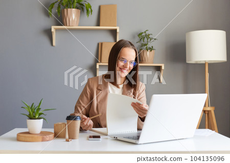 Portrait of good looking smiling attractive young adult woman with brown hair wearing beige jacket working online on laptop and with paper documents, expressing positive emotions. 104135096