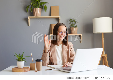 Indoor shot of attractive caucasian smiling friendly woman with brown hair wearing beige jacket working online on laptop, having video call, waving hand, saying hello or bye. 104135099