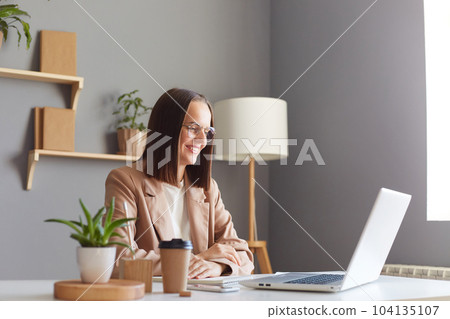 Profile portrait of charming adorable woman with brown hair wearing beige jacket working on laptop, looking at notebook display with toothy smile, having online meeting. Profile portrait of charming adorable woman with brown hair wearing beige jacket working on laptop, looking at notebook display with toothy smile, having online meeting. 104135107