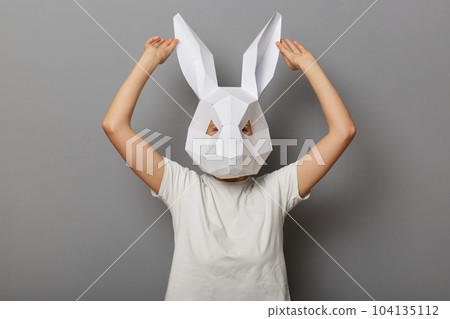 Indoor shot of funny anonymous female wearing rabbit mask and white t-shirt isolated over gray background, touching her paper ears, being in festive mood. 104135112