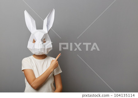Photo of unknown woman wearing white T-shirt and paper rabbit mask, pointing index finger aside at advertisement area, copy space for promotion, standing isolated over gray background. 104135114