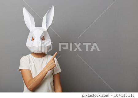 Horizontal shot of woman wearing white t shirt and paper rabbit mask standing isolated over gray background, standing pointing away at advertisement area, copy space. 104135115